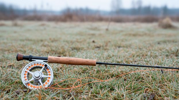 Caña de pescar a mosca apoyada sobre la hierba húmeda de un prado en invierno, con el carrete visible y la línea extendida.